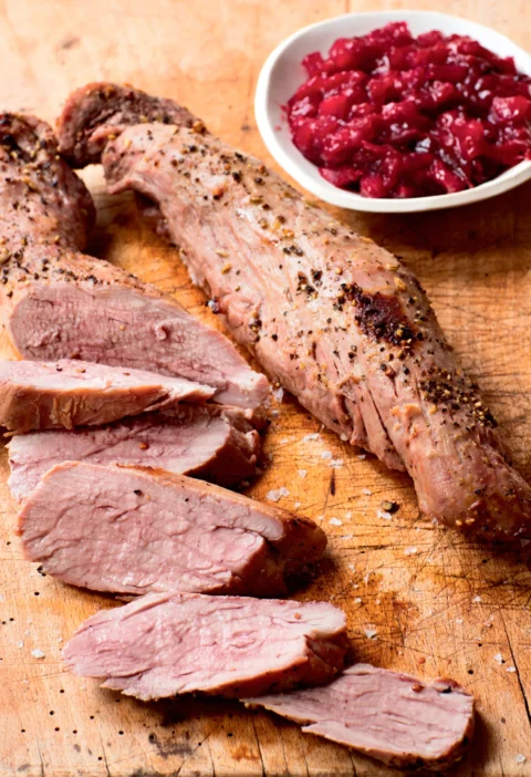 Sliced cider-roasted pork tenderloin on a wooden cutting board with a bowl of roasted plum chutney