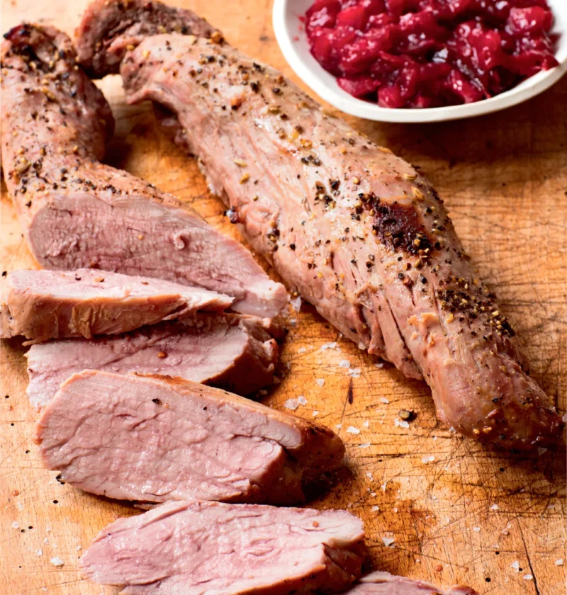 Sliced cider-roasted pork tenderloin on a wooden cutting board with a bowl of roasted plum chutney