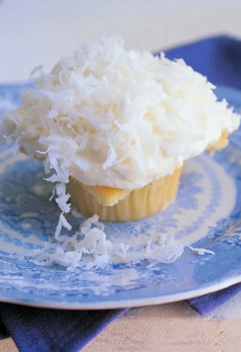 Ina Garten coconut cupcakes topped with cream cheese icing and shredded coconut on a blue and white plate