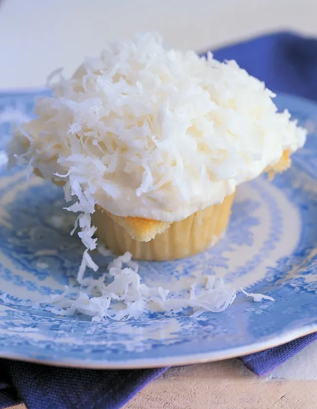 Ina Garten coconut cupcakes topped with cream cheese icing and shredded coconut on a blue and white plate