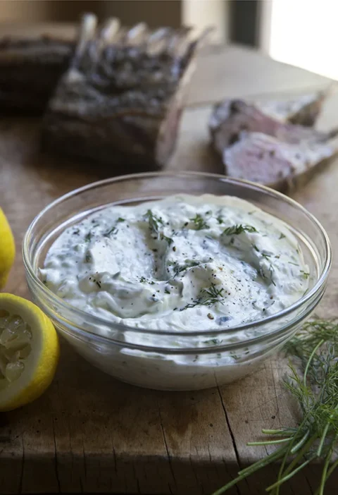 Ina Garten easy tzatziki in a glass bowl with fresh dill and lemon halves beside sliced lamb on a wooden board