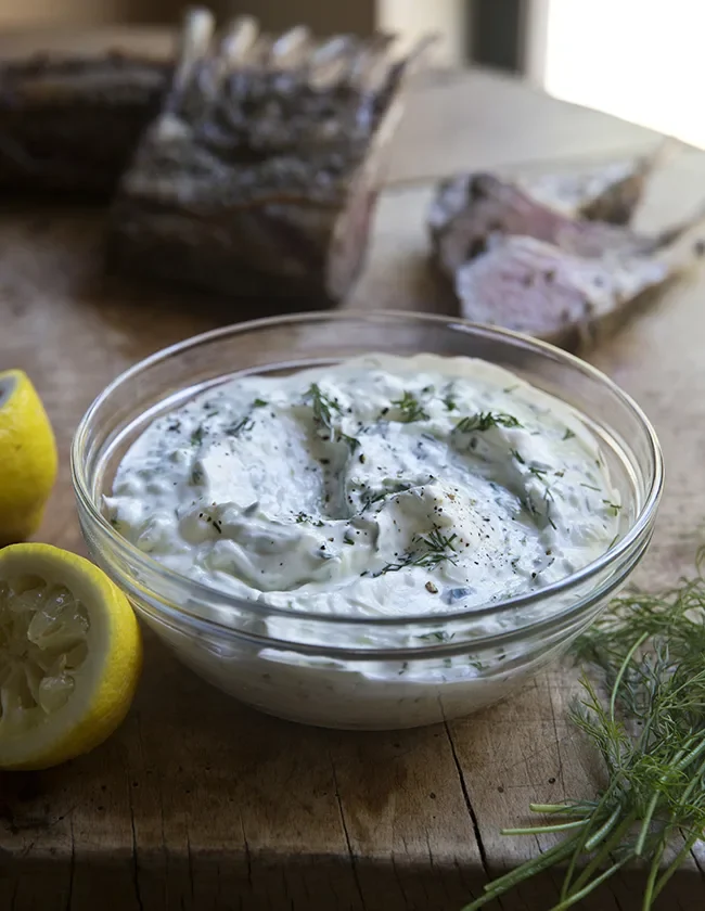 Ina Garten easy tzatziki in a glass bowl with fresh dill and lemon halves beside sliced lamb on a wooden board