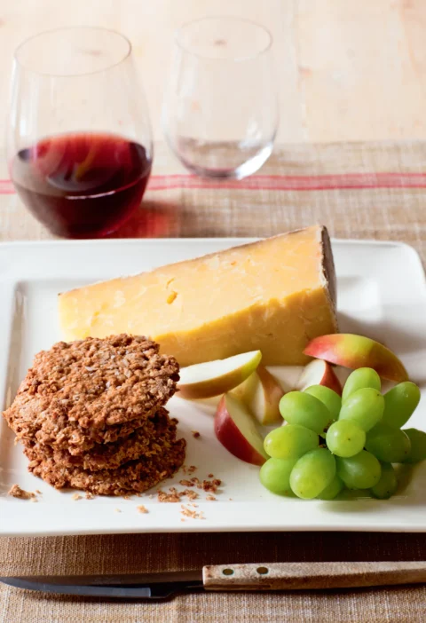 Ina Garten English oat crackers stacked on a white plate next to a wedge of Cheddar, apple slices and green grapes