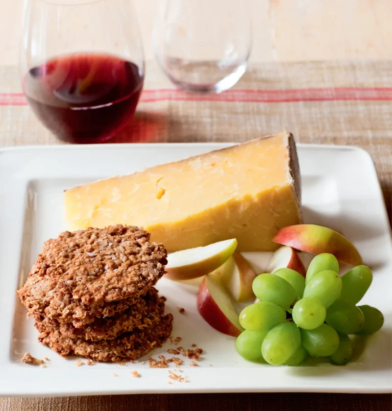 Ina Garten English oat crackers stacked on a white plate next to a wedge of Cheddar, apple slices and green grapes