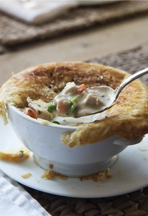 Ina Garten French chicken pot pie in a white bowl with golden puff pastry crust and a spoon lifting creamy chicken, carrots and peas