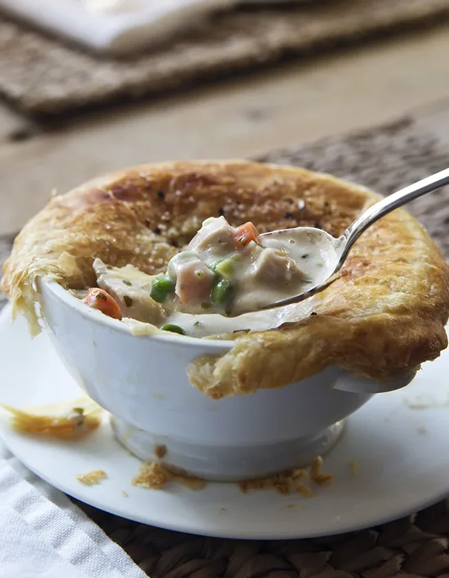 Ina Garten French chicken pot pie in a white bowl with golden puff pastry crust and a spoon lifting creamy chicken, carrots and peas