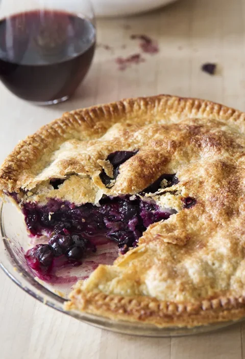 Ina Garten fresh blueberry pie with a golden double crust and a slice cut out showing the jammy blueberry filling, with a glass of red wine behind it