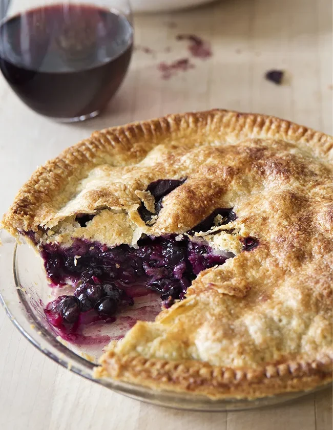Ina Garten fresh blueberry pie with a golden double crust and a slice cut out showing the jammy blueberry filling, with a glass of red wine behind it