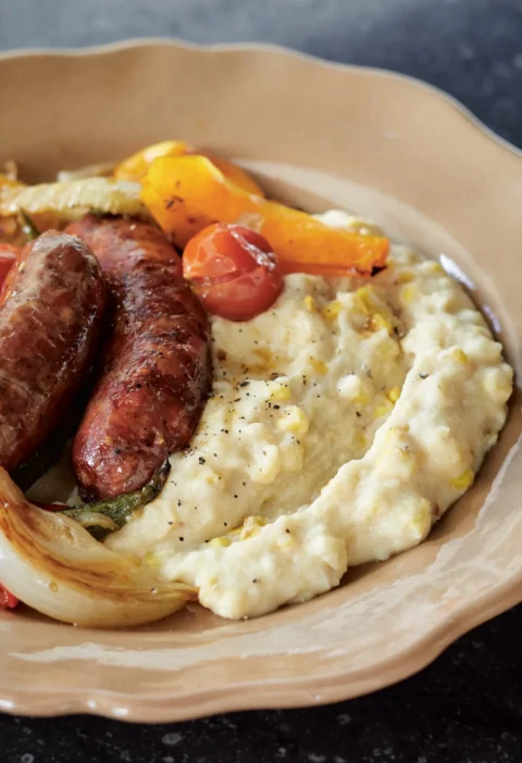 Ina Garten fresh corn polenta served in a shallow bowl next to roasted sausages, peppers and onions