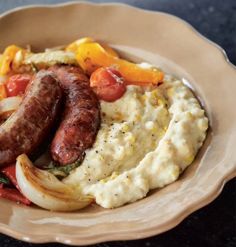 Ina Garten fresh corn polenta served in a shallow bowl next to roasted sausages, peppers and onions
