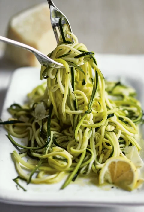 Ina Garten fresh zucchini with lemon & mint, long julienned zucchini strands twirled on a fork over a white dish with lemon wedges and Parmesan