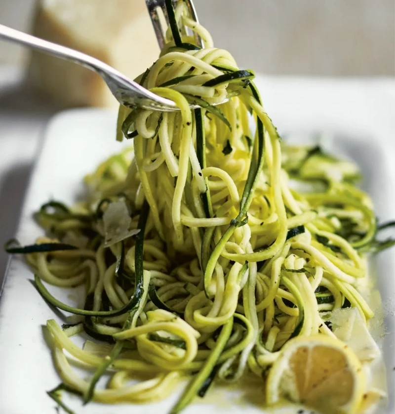 Ina Garten fresh zucchini with lemon & mint, long julienned zucchini strands twirled on a fork over a white dish with lemon wedges and Parmesan
