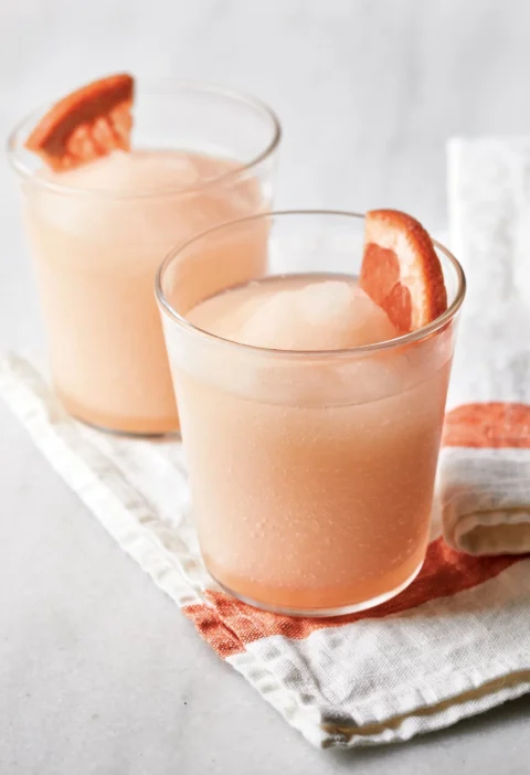 Two glasses of Ina Garten frozen palomas, pink grapefruit slushies garnished with ruby red grapefruit wedges on a striped linen napkin