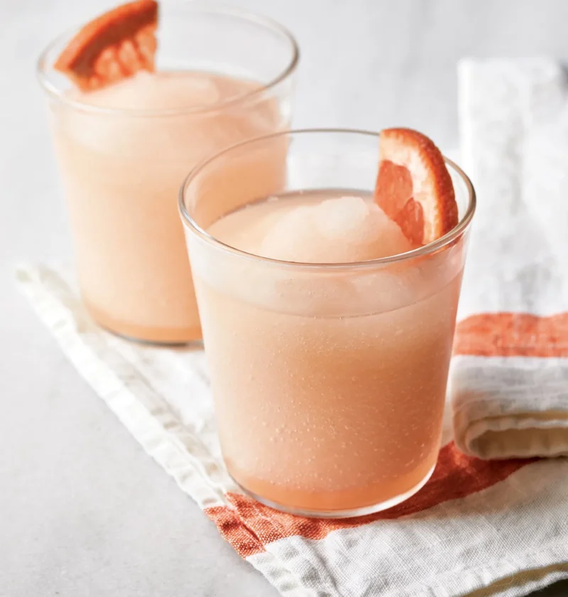 Two glasses of Ina Garten frozen palomas, pink grapefruit slushies garnished with ruby red grapefruit wedges on a striped linen napkin