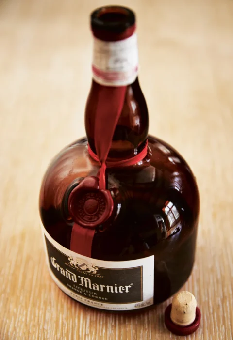 Open bottle of Grand Marnier with red ribbon and wax seal next to its cork on a light wood surface, the liqueur used in Ina Garten Grand Marnier pastry cream