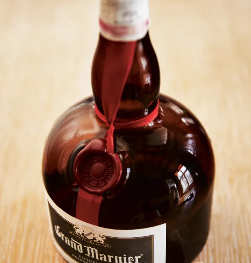 Open bottle of Grand Marnier with red ribbon and wax seal next to its cork on a light wood surface, the liqueur used in Ina Garten Grand Marnier pastry cream