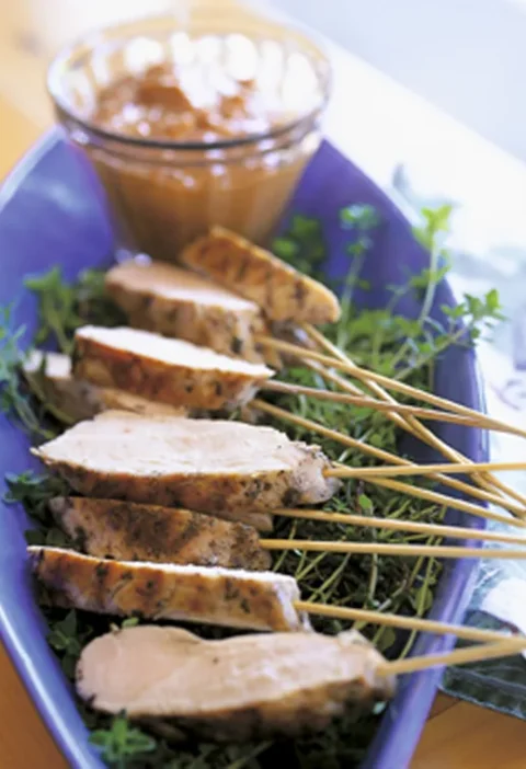 Ina Garten grilled lemon chicken sliced on wooden skewers over fresh thyme on a blue platter with a small bowl of satay dip