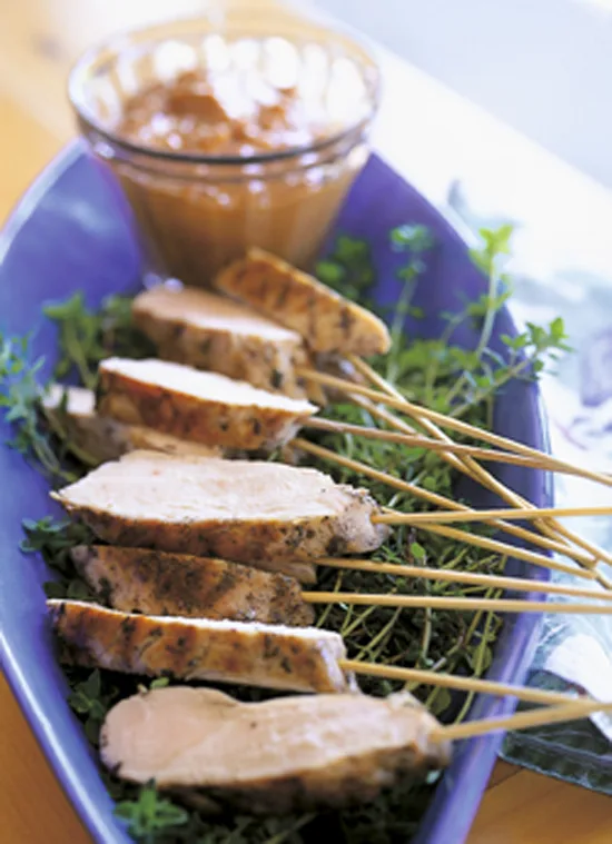 Ina Garten grilled lemon chicken sliced on wooden skewers over fresh thyme on a blue platter with a small bowl of satay dip