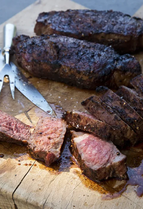 Ina Garten grilled New York strip steaks with a dark spice crust, sliced medium rare on a wooden cutting board with a carving fork