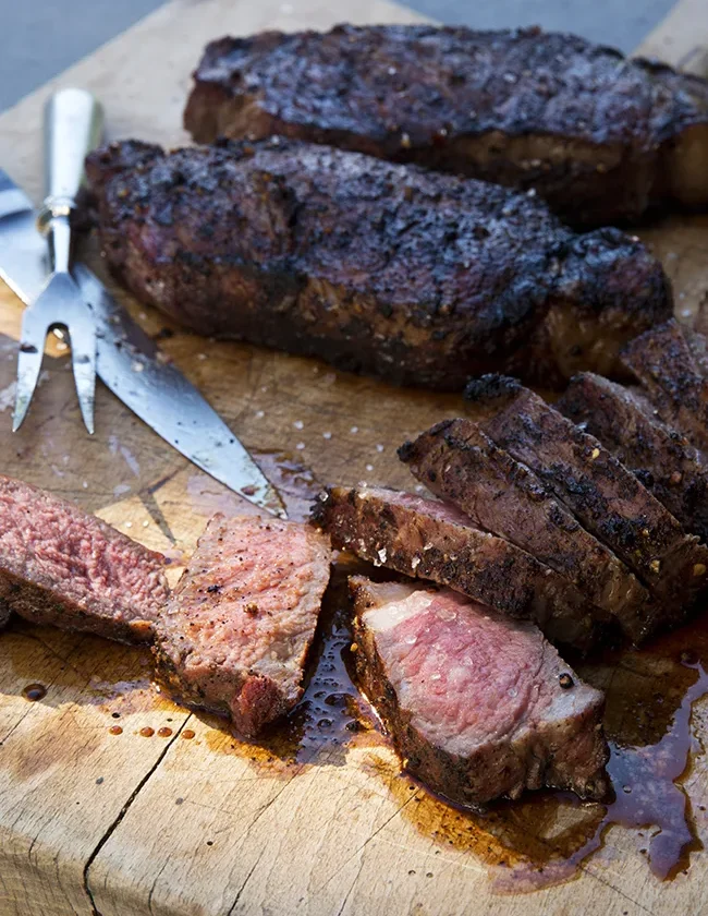 Ina Garten grilled New York strip steaks with a dark spice crust, sliced medium rare on a wooden cutting board with a carving fork