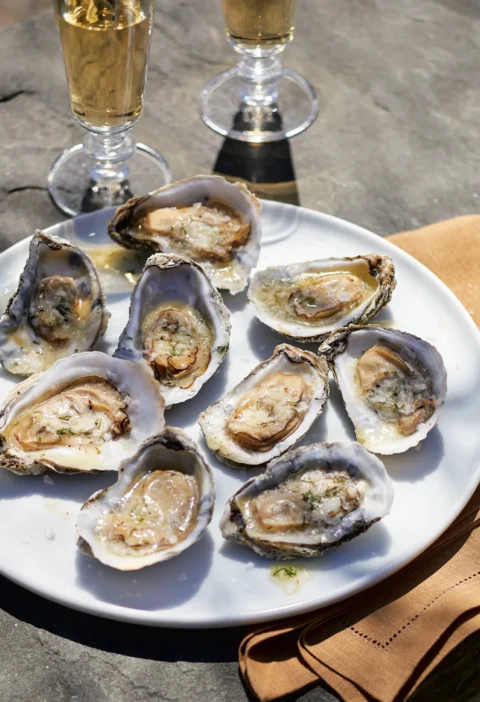 Ina Garten grilled oysters with lemon dill butter on the half shell on a white plate with glasses of white wine