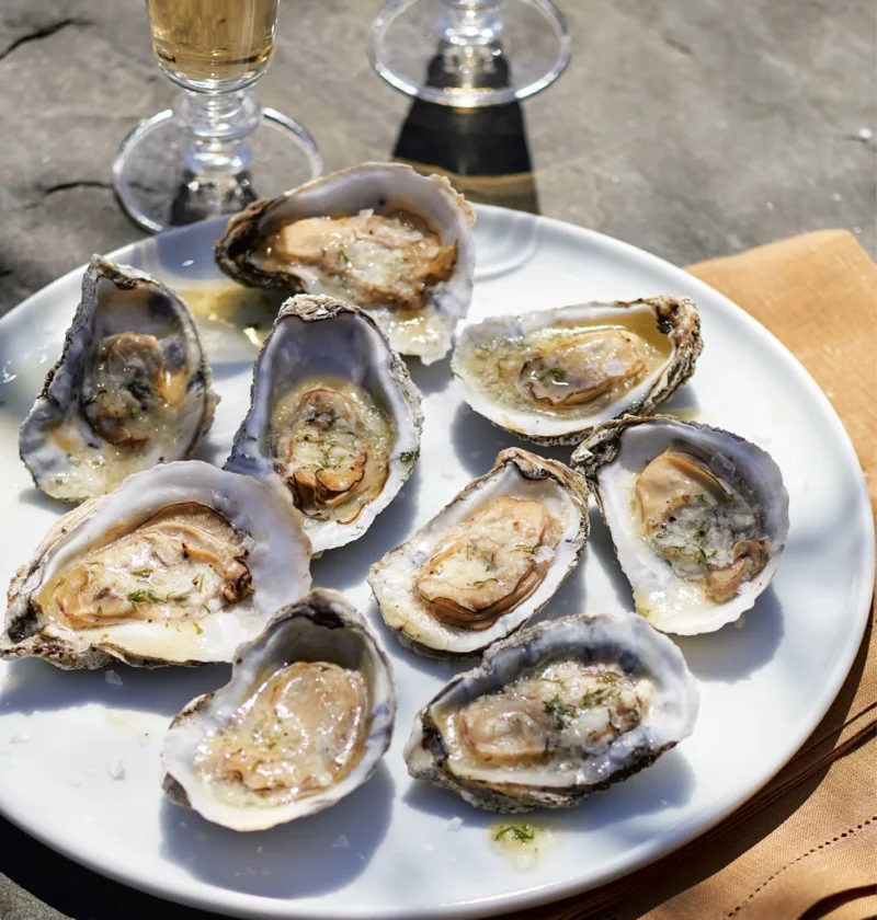 Ina Garten grilled oysters with lemon dill butter on the half shell on a white plate with glasses of white wine