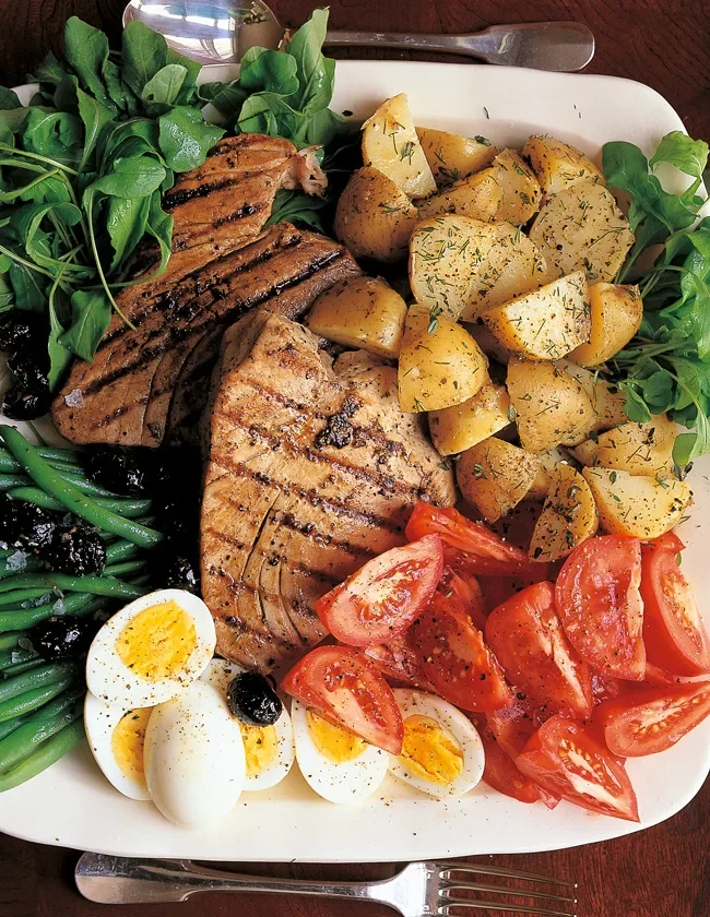 Ina Garten grilled tuna niçoise platter with seared tuna steaks, potato salad, tomato wedges, halved eggs, haricots verts and olives on a white platter