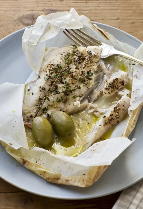 Ina Garten herb-roasted fish in an opened parchment packet with fresh thyme sprigs, green olives and lemon olive oil juices on a blue plate