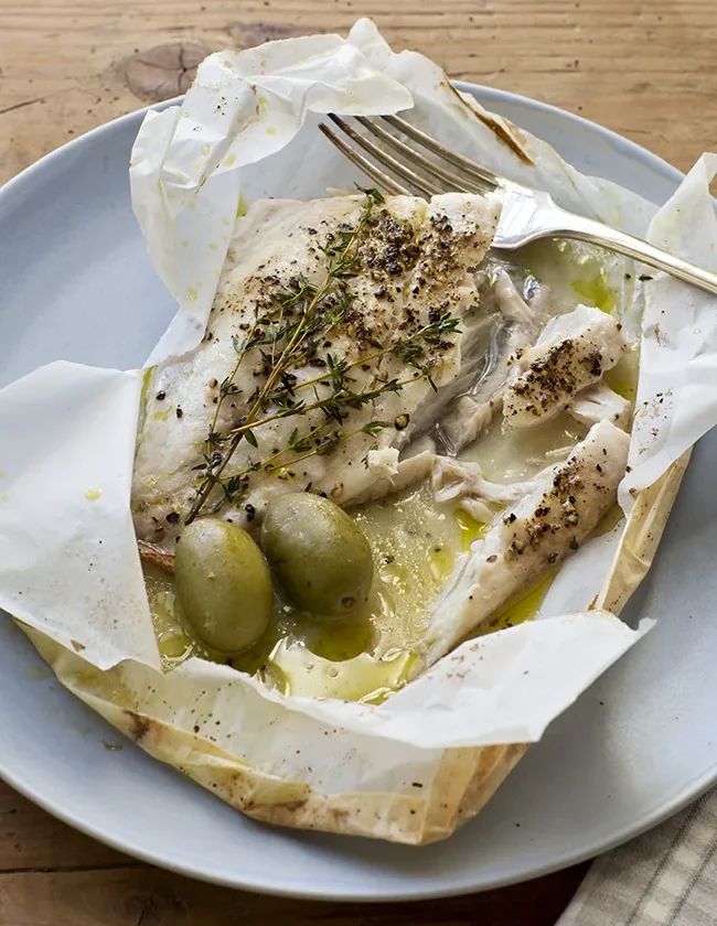 Ina Garten herb-roasted fish in an opened parchment packet with fresh thyme sprigs, green olives and lemon olive oil juices on a blue plate