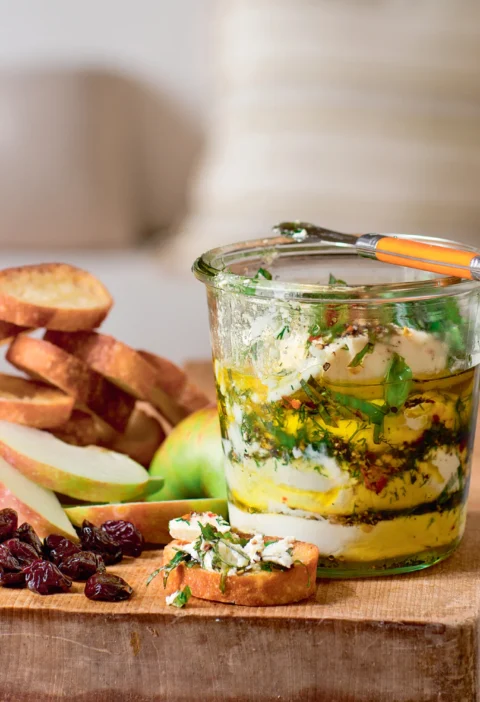 Ina Garten herbed goat cheese layered in a glass jar with olive oil, dill and basil, served with crostini, apple slices and dried cranberries