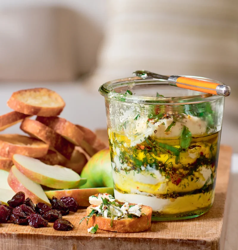 Ina Garten herbed goat cheese layered in a glass jar with olive oil, dill and basil, served with crostini, apple slices and dried cranberries