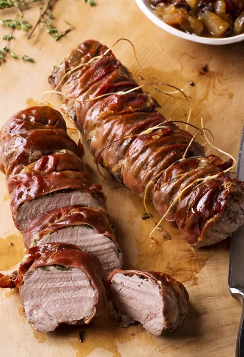 Ina Garten herbed pork tenderloins with apple chutney, prosciutto-wrapped and sliced on a wooden cutting board