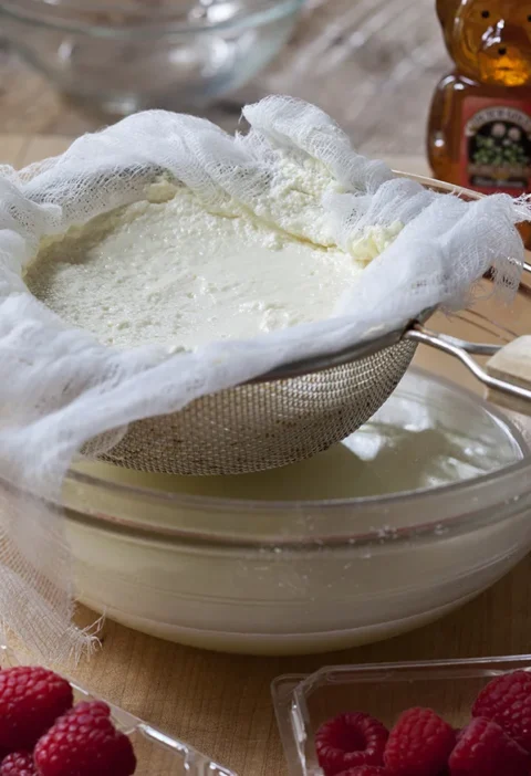Ina Garten homemade ricotta draining in a cheesecloth-lined sieve over a glass bowl with fresh raspberries nearby