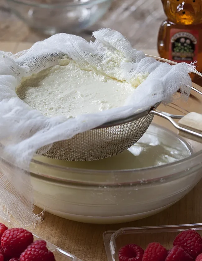 Ina Garten homemade ricotta draining in a cheesecloth-lined sieve over a glass bowl with fresh raspberries nearby