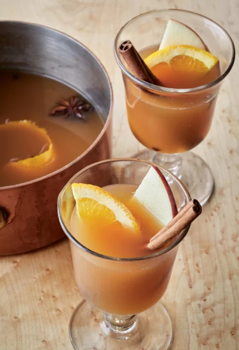 Ina Garten hot spiced apple cider in glasses with orange slices, apple slices and cinnamon sticks next to a copper saucepan