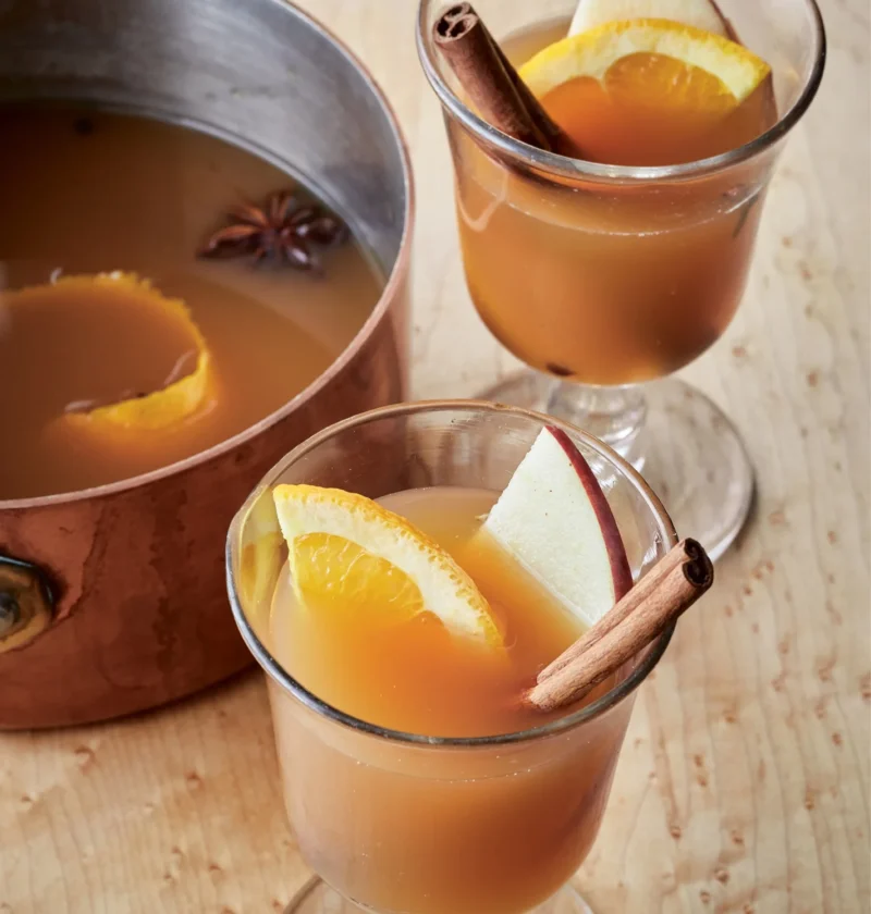Ina Garten hot spiced apple cider in glasses with orange slices, apple slices and cinnamon sticks next to a copper saucepan