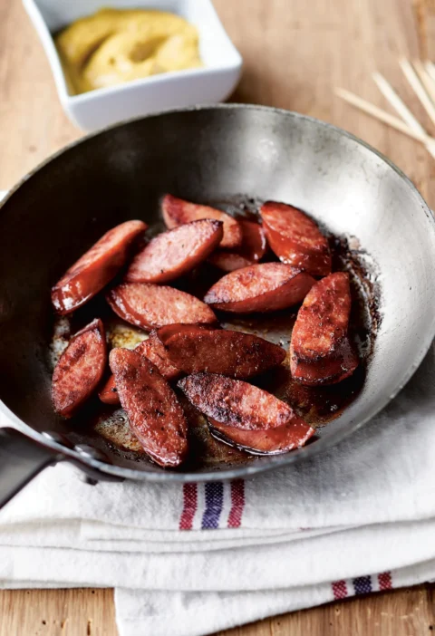 Ina Garten kielbasa with mustard dip: seared kielbasa slices in a skillet with a bowl of mustard sauce and wooden skewers