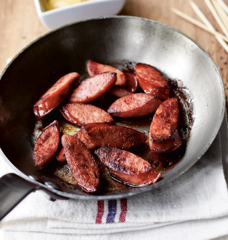 Ina Garten kielbasa with mustard dip: seared kielbasa slices in a skillet with a bowl of mustard sauce and wooden skewers