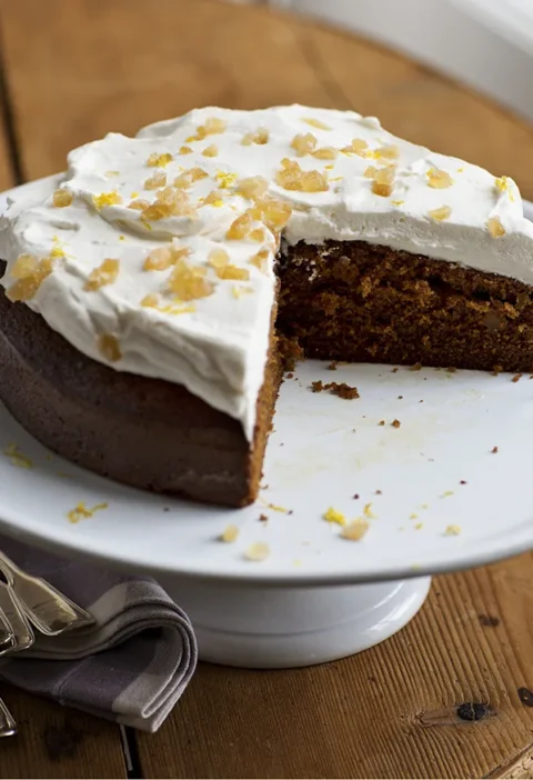Ina Garten lemon ginger molasses cake topped with whipped cream and crystallized ginger on a white cake stand with a slice removed