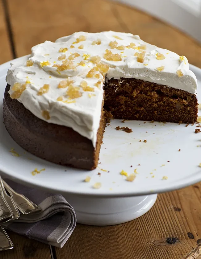 Ina Garten lemon ginger molasses cake topped with whipped cream and crystallized ginger on a white cake stand with a slice removed
