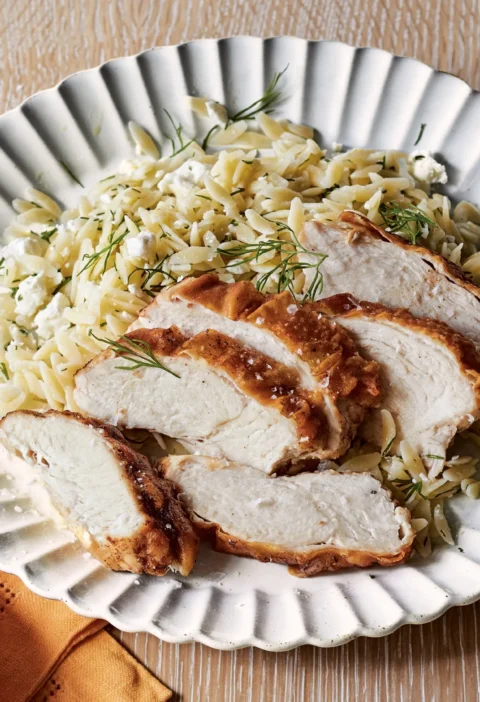 Ina Garten lemon orzo with feta topped with fresh dill and served with sliced chicken breast on a white scalloped plate