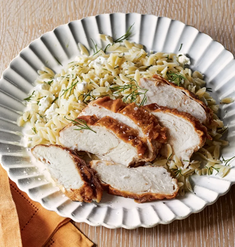 Ina Garten lemon orzo with feta topped with fresh dill and served with sliced chicken breast on a white scalloped plate