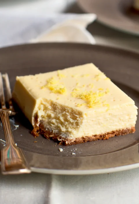Ina Garten limoncello ricotta cheesecake square with a bite taken out, topped with lemon zest on a graham cracker crust