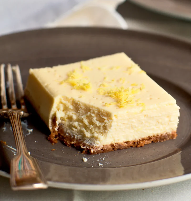 Ina Garten limoncello ricotta cheesecake square with a bite taken out, topped with lemon zest on a graham cracker crust