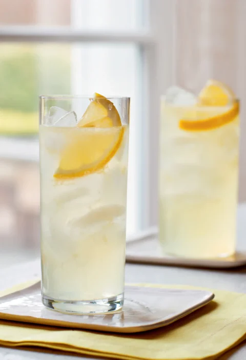 Two tall glasses of Ina Garten limoncello vodka collins with ice and lemon slices on a yellow napkin