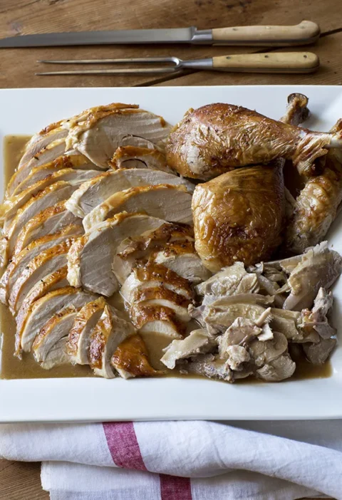 Ina Garten make-ahead roast turkey sliced on a white platter over gravy with carving knife and fork