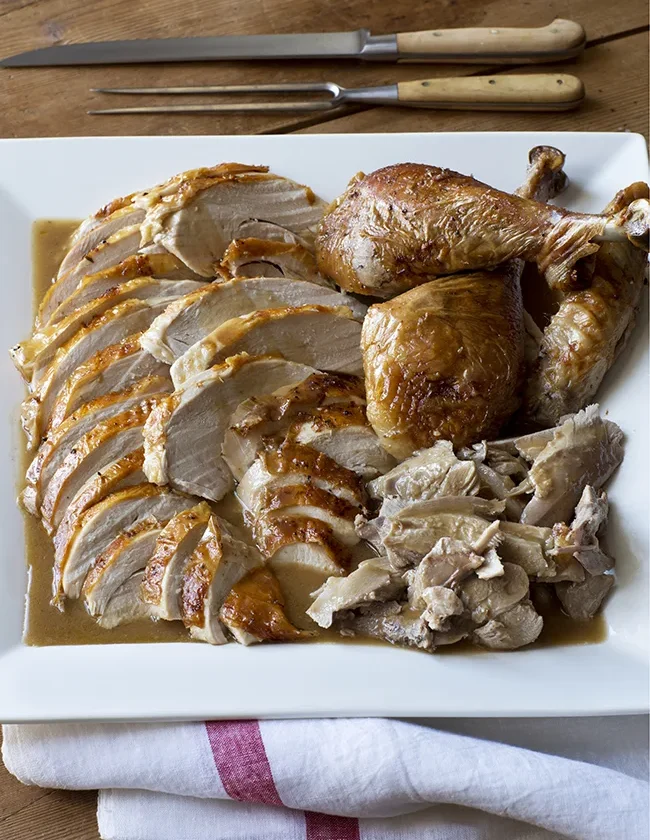 Ina Garten make-ahead roast turkey sliced on a white platter over gravy with carving knife and fork