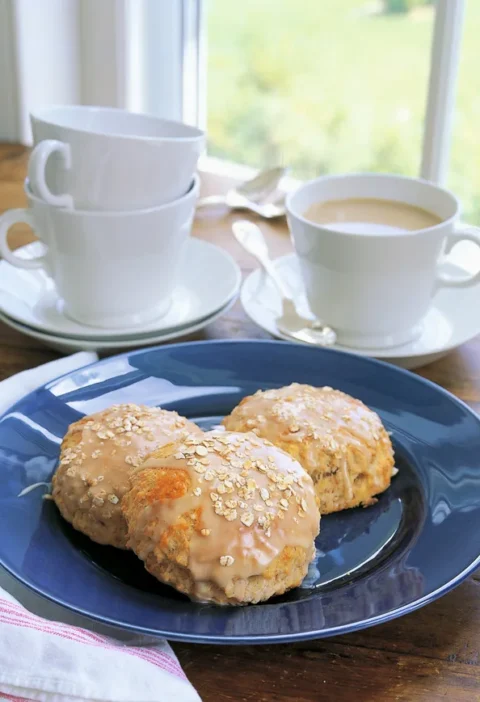 Ina Garten maple-oatmeal scones with maple glaze and oats on a blue plate next to a cup of coffee