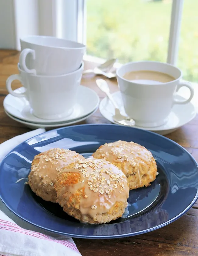 Ina Garten maple-oatmeal scones with maple glaze and oats on a blue plate next to a cup of coffee
