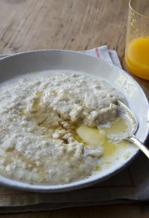 Ina Garten maple vanilla cream of wheat in a gray bowl with melting butter and maple syrup, next to a glass of orange juice
